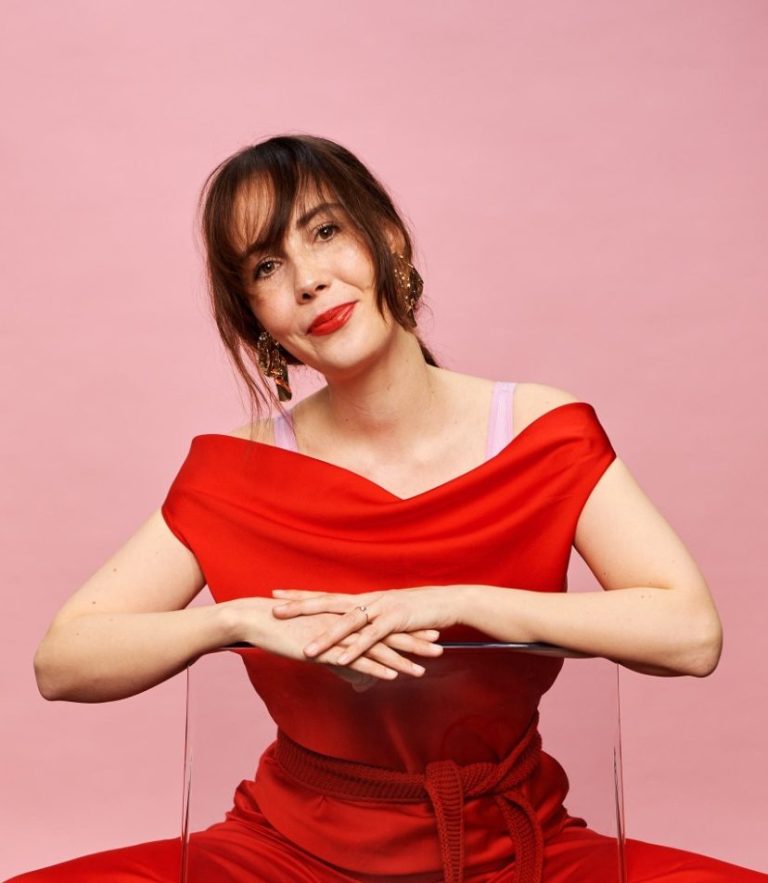 Frau in rotem Kleid mit offener Blende, lächelt vor pinkem Hintergrund.