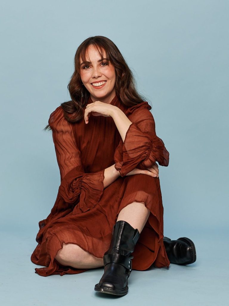 Eine lächelnde Person sitzt in einem braunen Kleid und schwarzen Stiefeln vor einem blauen Hintergrund.