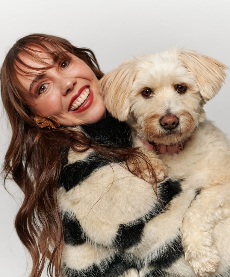 Frau mit lockigem, braunem Haar umarmt einen Hund mit hellbeigem Fell.