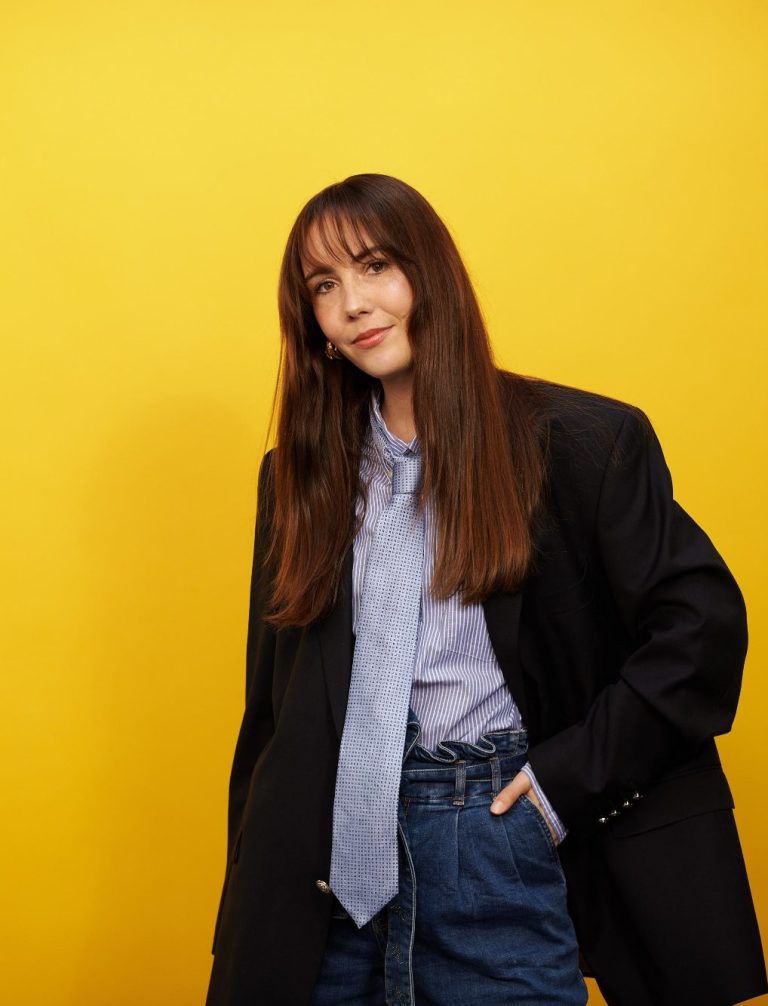 Frau mit langen Haaren in einem schwarzen Blazer vor gelbem Hintergrund.